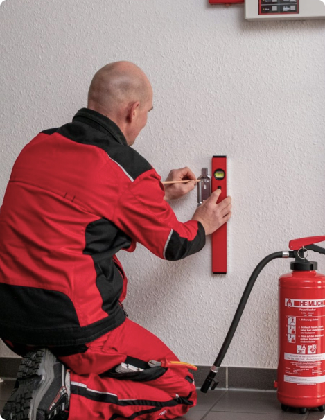 Ein Arbeiter in Schutzkleidung nimmt mit Hilfe einer Wasserwaage und eines Bleistifts Markierungen an der Wand neben dem bereits montierten Feuerlöscher vor.