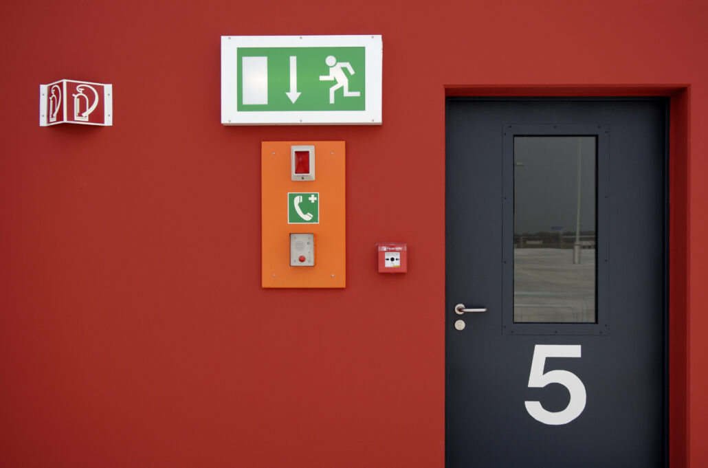 Ein grünes Notausgangsschild, eine SOS-Sprechanlage, ein Feueralarmknopf, ein Piktogramm für einen Feuerlöscher und eine schwarze Tür mit der Nummer 5 an einer roten Wand.
