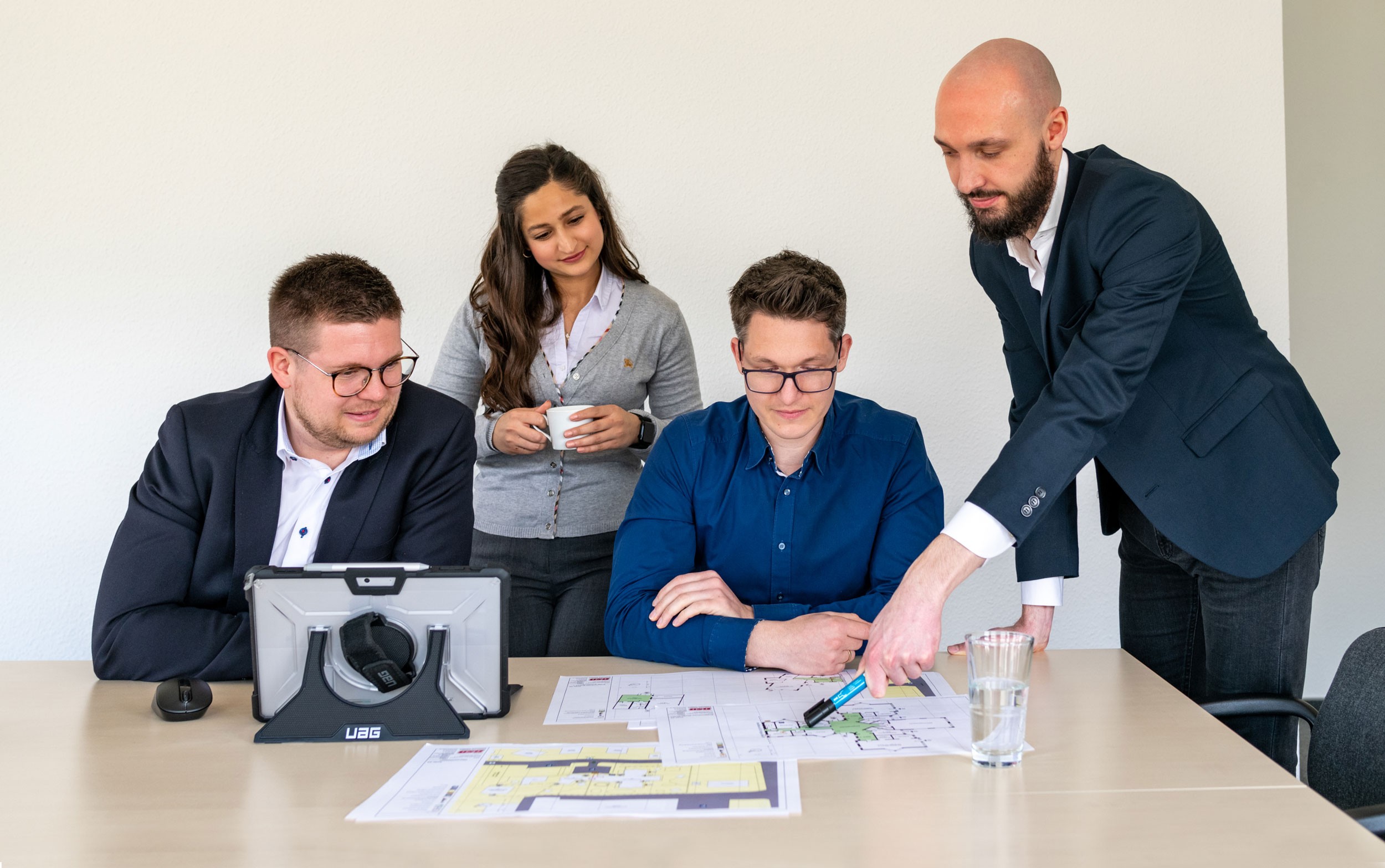 Eine Gruppe von vier Mitarbeitern der BSB Kröger arbeitet an einem Tisch an Evakuierungsplänen. Zwei sitzen und konzentrieren sich auf die Pläne, während die anderen stehen, sich austauschen und Ideen diskutieren.