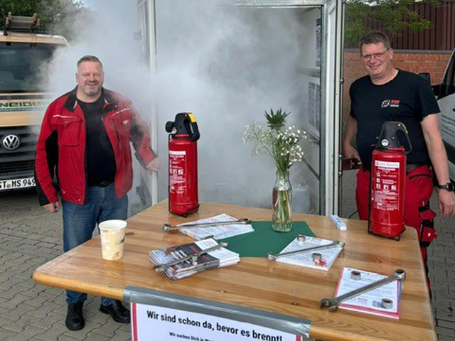 Zwei Mitarbeiter von BSB Kröger stehen auf einem Straßenmarkt zu beiden Seiten eines Tisches mit Feuerlöschern und führen die Funktionsweise der Geräte vor